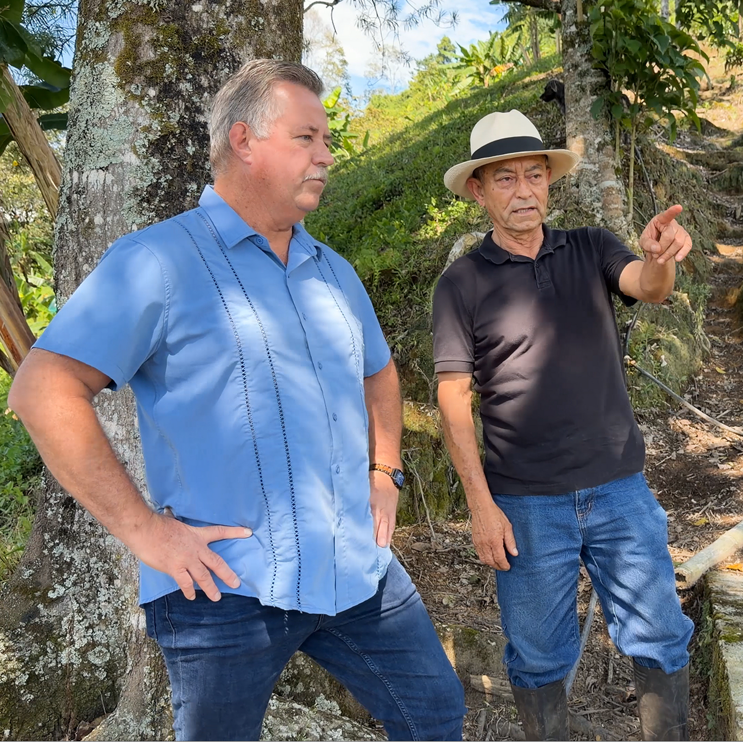 Two men standing outdoors in a natural setting, one pointing towards something off-camera.